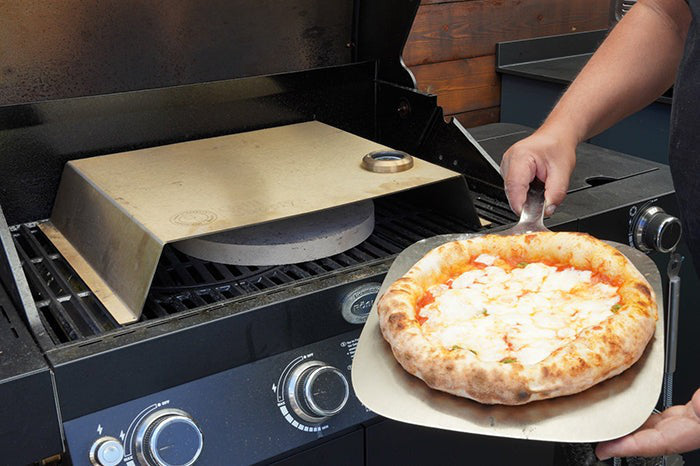 Eine Pizza wird auf einem Gasgrill mit Hilfe eines Pizzastein zubereitet.