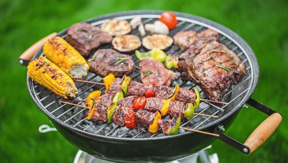 Verschiedene Fleischsorten auf einem Kugelgrill, zusammen mit Mais, Tomaten und weiterem Gemüße